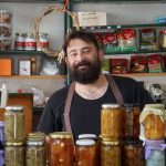 Pequeno comerciante atrás de prateleiras com conservas artesanais em seu estabelecimento.