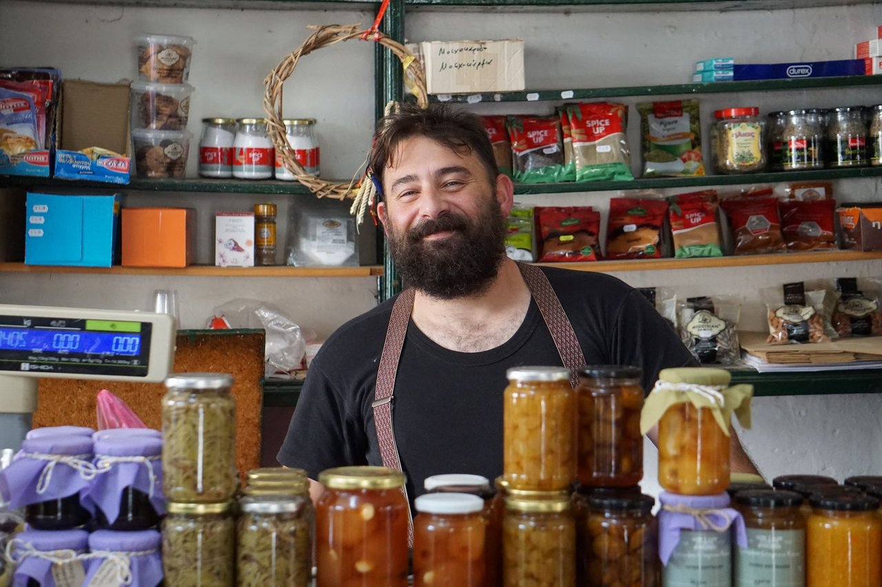Pequeno comerciante atrás de prateleiras com conservas artesanais em seu estabelecimento.